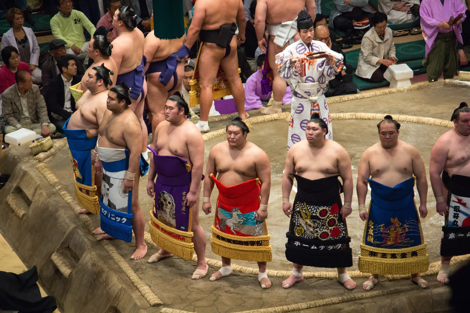 Un cerchio di lottatori di sumo in colorati grembiuli cerimoniali in piedi su una piattaforma di argilla in un'arena, con il pubblico tutt'intorno