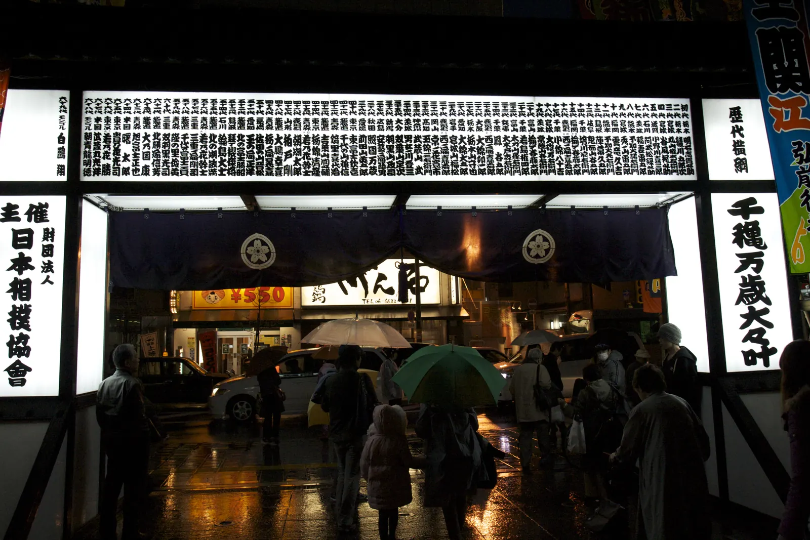 L'ingresso illuminato di una sede di sumo di notte, con alti stendardi di calligrafia nera su bianco e persone in piedi sotto gli ombrelli