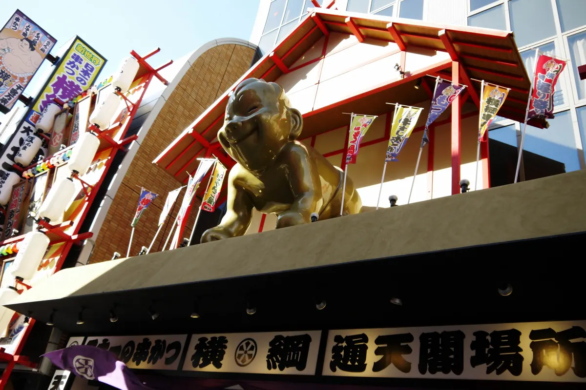 Eine vergoldete sitzende Billiken-Figur mit spitzem Kopf und breitem Lächeln auf einem rot-goldenen Restaurantschild in Osaka