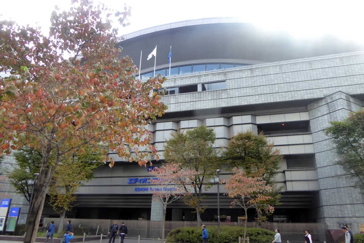 The grey concrete exterior of EDION Arena Osaka behind trees with autumn leaves, with people walking on the pavement in front