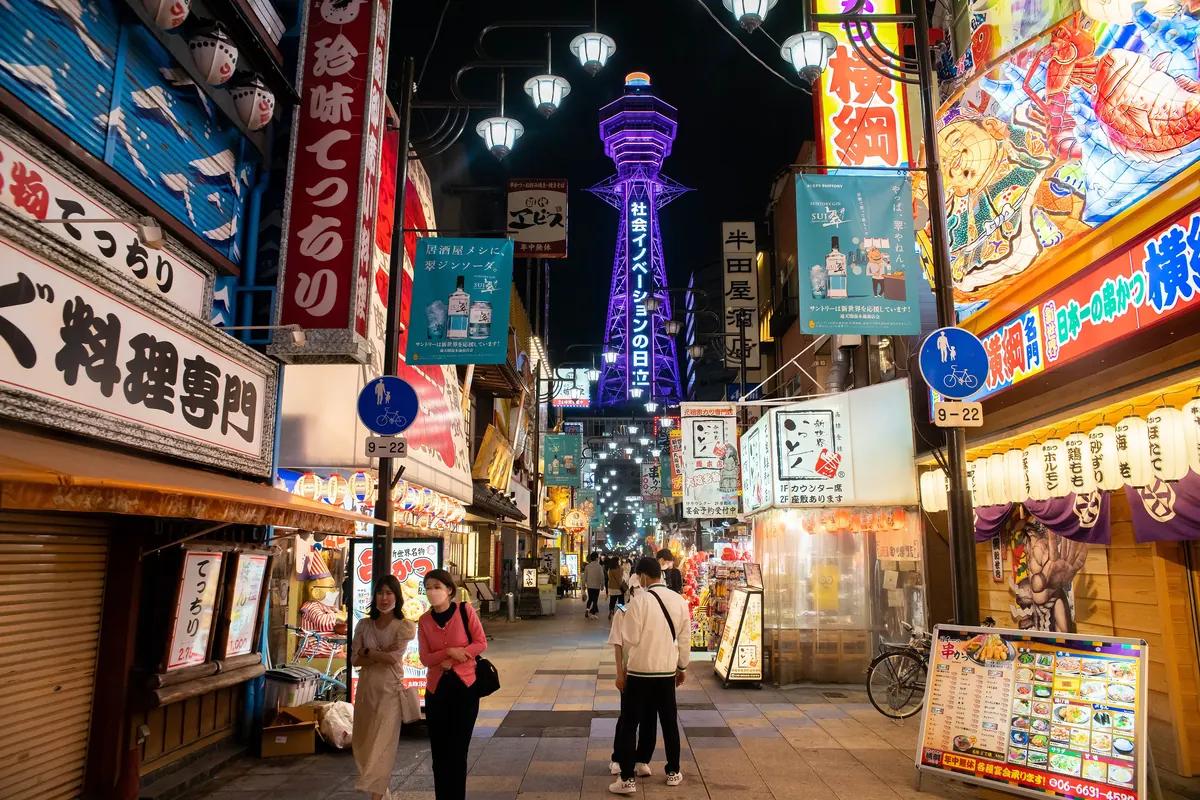 Ein hoch aufragender, beleuchteter Gitterturm, der nachts in Shinsekai purpurfarben über einer Straße voller Neonreklamen und Lampions erstrahlt
