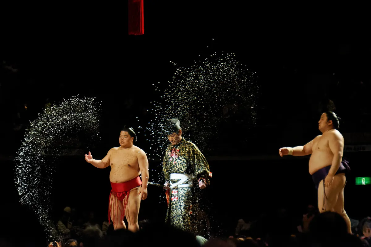 Sumo-Ringer in einer dunklen Arena, ein Sprühregen aus weißem Salz in der Luft um einen Kampfrichter in zeremonieller Kleidung