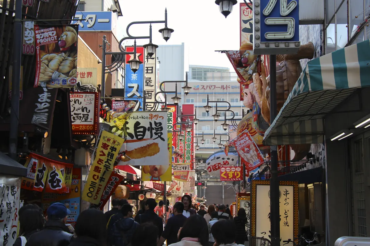 Eine belebte, enge Straße in Osaka, voller Ladenschilder, Laternen und Banner von Food-Ständen über den Köpfen der Passanten