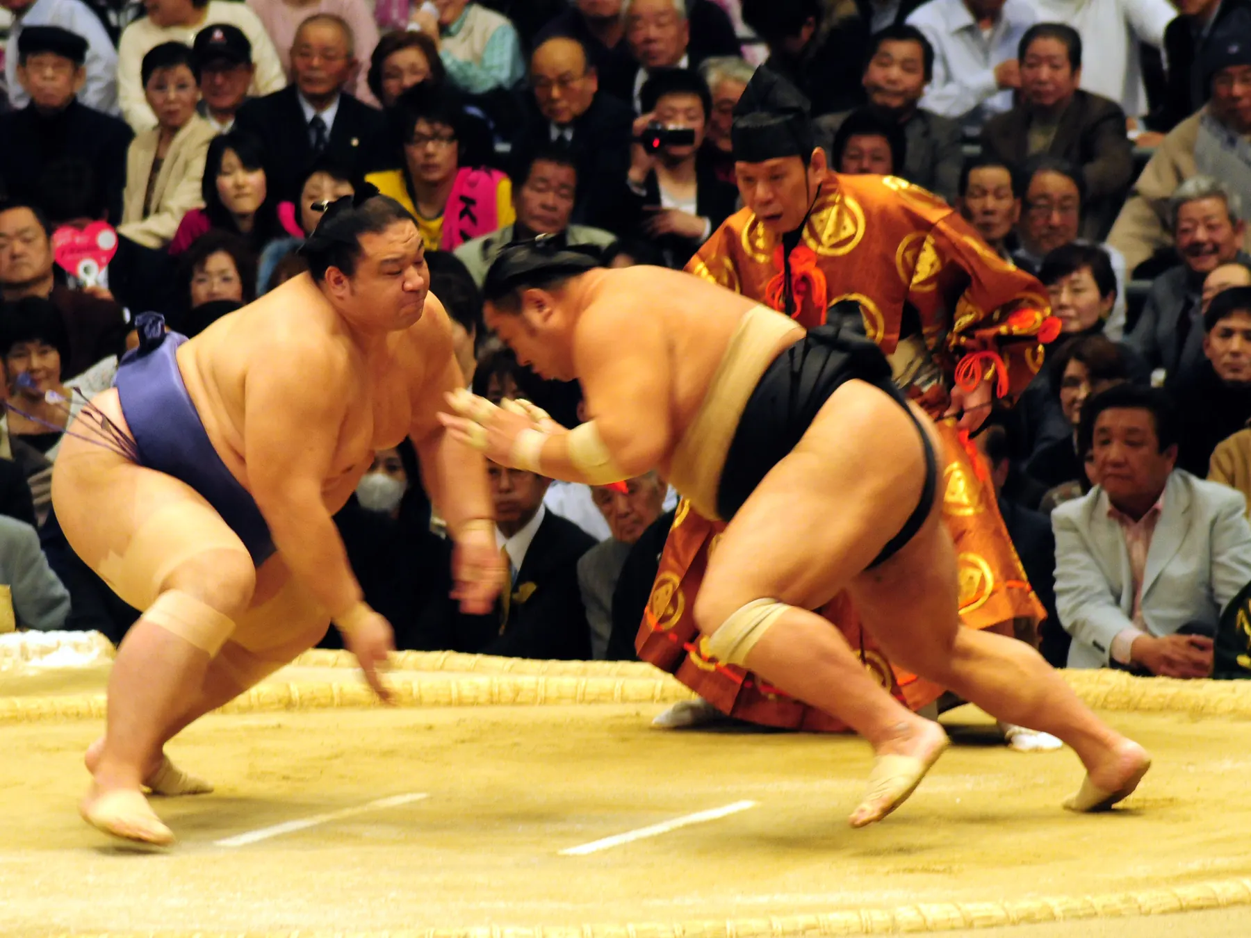 Zwei Sumo-Ringer, die zu Beginn eines Kampfes in einem Ring aus Strohballen aufeinander zustürmen, mit einem Schiedsrichter in orangefarbener Zeremonienkleidung hinter ihnen und einem sitzenden Publikum dahinter